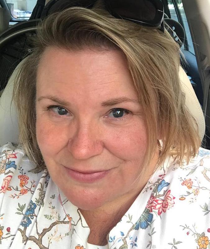 Maria, solbränd med solglasögon i pannan, iklädd ljus mönstrad topp, sitter i förarsäte i bil, i bakgrunden syns rullstolen baksätet.