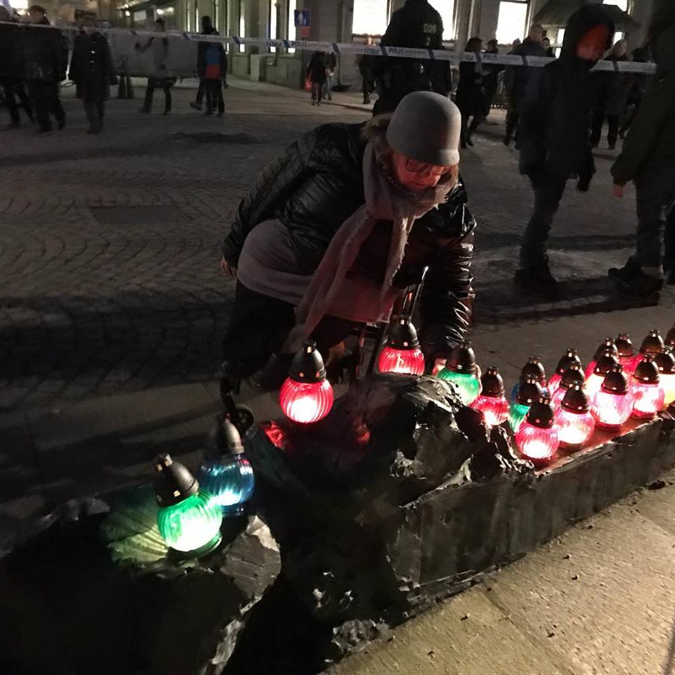 Jag, klädd i svart och grått, ställer ned en grön lysande glaslykta bland många andra, på de svarta stenkonstverken på torget, tidig kväll, i bakgrunden syns polisavspärrning.