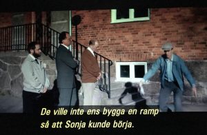 Filmruta. Skolgård, entrétrappa. Ove måttar med armarna hur en ramp skulle gå, tre män står med armarna i kors och tittar på. Speakerröst/textremsa ”De ville inte ens bygga en ramp så att Sonja kunde börja.”