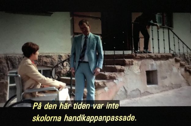 Filmruta. Skolgård, trappa till huvudentrén, nedanför trappan sitter Sonja i rullstol, hon pratar med man som står. Speakerröst/textremsa ”På den tiden var inte skolorna handikappanpassade.”
