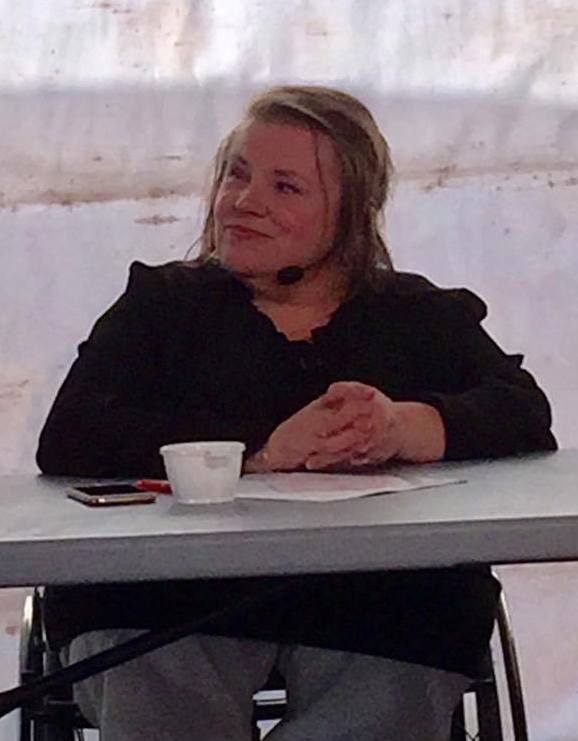 Jag klädd i svart bakom bord i panel i Almedalen, mikrofon i headset, telefon, papper och plastmugg framför mig på bordet.